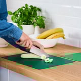 Personne coupant la banane sur un tapis de découpe vert flexible, protégeant le comptoir en bois.