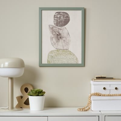 Des œuvres d’art abstraites encadrées sont accrochées à un mur beige au-dessus d’une armoire blanche. Une lampe blanche et une petite plante en pot se trouvent sur l’armoire.