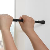 La personne installe la tringle à rideau FJÄDRANDE, en la tendant entre le cadre de la fenêtre et le mur.