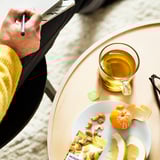 Personne écrivant dans un cahier à côté d’une table avec du thé, des fruits et des noix de cajou.