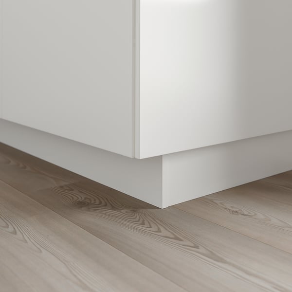 Un gros plan du socle d’armoires de cuisine blanches, fixé au-dessus de planches de bois clair. Il dissimule l’espace entre le sol et l’armoire basse.