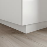 Un gros plan du socle d’armoires de cuisine blanches, fixé au-dessus de planches de bois clair. Il dissimule l’espace entre le sol et l’armoire basse.
