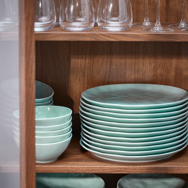 Étagère en bois contenant des piles de bols et assiettes en porcelaine verte et blanche, avec des verres transparents sur le dessus.