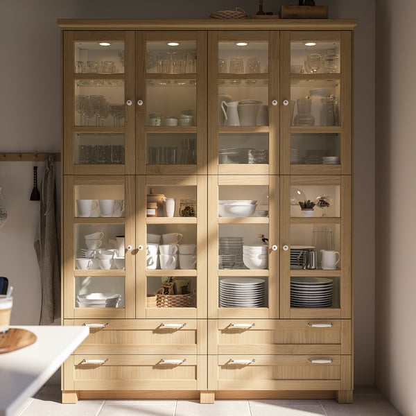 Armoire de cuisine en bois, design FORSBACKA, couleur brun clair, portes en verre, plusieurs étagères, vaisselle bien organisée, tiroir en bas.