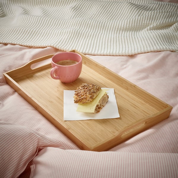 Plateau de petit-déjeuner en bois sur un lit avec des draps roses, contenant une tasse de café et un sandwich, offrant une expérience de petit-déjeuner confortable.