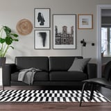 Salon moderne avec canapé FULLERÖ noir, coussins gris, tapis noir et blanc. En face d’une chaise en cuir gris. Œuvre d’art au-dessus du canapé.