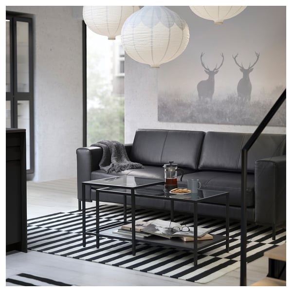 Salon moderne avec canapé en cuir FULLERÖ noir, table basse en verre, tapis rayé et lanternes suspendues.