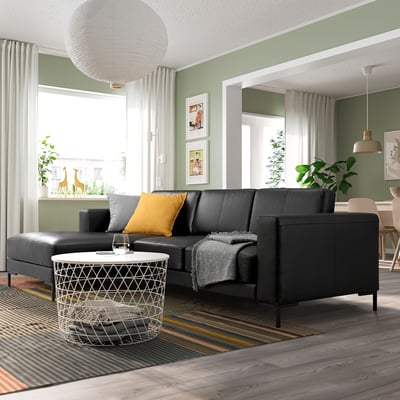 Salon moderne avec sectionnel FULLERÖ noir, oreiller jaune, table basse blanche, murs verts, sols rayés et grandes fenêtres.