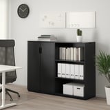 Une unité de rangement de bureau noire GALANT avec des étagères fermées et ouvertes contenant des livres, des classeurs et une plante. À côté, une chaise blanche est visible.