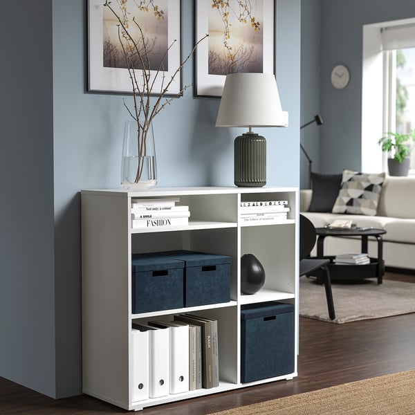 Boîte de rangement GJÄTTA en velours blanc sur étagère, compacte et fonctionnelle, sous lampe parmi d’autres boîtes et livres.
