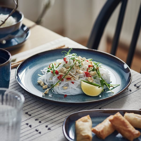 Assiette émaillée bleue avec plat de nouilles et quartier de citron vert, posée sur une table en bois avec des assiettes supplémentaires et un verre.