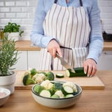 Personne coupant des courgettes dans un bol de légumes sur une planche à découper en bois avec un plat à four à proximité.