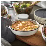 Une main dans un gant de cuisine gris tient un plat en céramique bleue avec des lasagnes, entouré de bols et d’une assiette avec des tomates et des herbes sur une planche de bois.