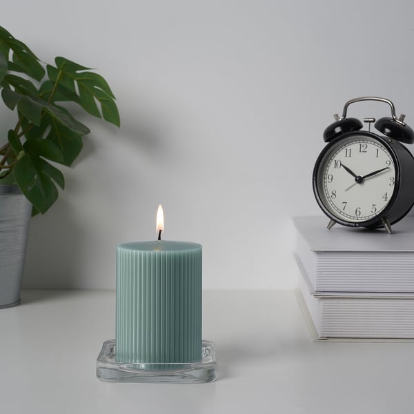 Une bougie verte allumée se trouve sur un support en verre à côté d’un réveil et d’une pile de livres.
