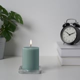 Une bougie verte allumée se trouve sur un support en verre à côté d’un réveil et d’une pile de livres.