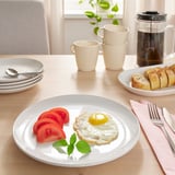 Une table de petit-déjeuner avec de la vaisselle blanche, avec un œuf au plat, des tranches de tomates, une fourchette, un couteau et des tasses à café.