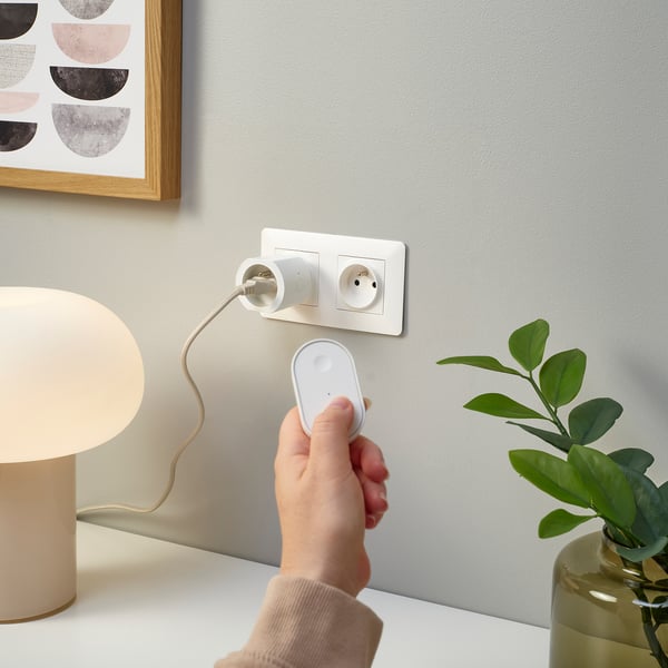 Une main utilise une télécommande pour contrôler une lampe branchée sur une prise intelligente. À côté d’une plante d’intérieur et de l’art mural.