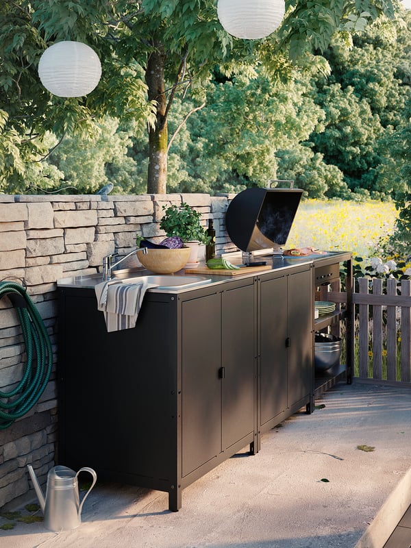 Cuisine extérieure avec gril noir, évier et comptoir. Des lanternes suspendues, des plantes en pot et un tuyau d’arrosage. Idéal pour les barbecues.