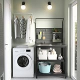 Petite unité de buanderie en métal noir avec évier et rangement, s’adapte parfaitement à côté de la machine à laver blanche, contenant des vêtements pliés et des fournitures de lessive.