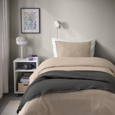 Lit douillet avec couette beige, couverture grise, applique murale et table de chevet.