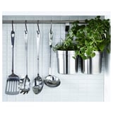 Une tringle de cuisine avec des cuillères métalliques, une louche, une spatule et des herbes dans des pots.