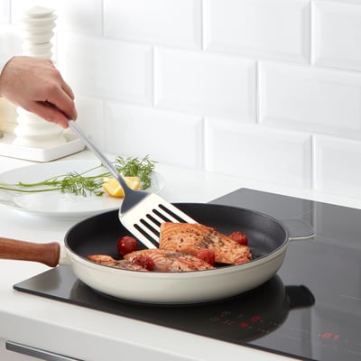 Une main tient une spatule avec du poisson cuit dans une poêle blanche sur une cuisinière. Une assiette aux herbes et au citron se trouve à proximité.