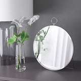 Un miroir rond sur un support à côté d’un vase transparent avec des fleurs blanches. Le miroir peut être suspendu ou debout.