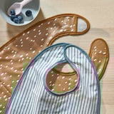 Deux bavoirs pour bébé à motifs sur une surface en bois. L’un avec des rayures, l’autre avec des pois. Les façades sont imperméables, les dos sont en coton doux.