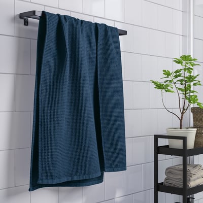 La serviette gaufrée bleue GULLDYNA est suspendue sur un porte-serviettes, près de petites plantes et serviettes pliées sur un support dans une salle de bain carrelée.