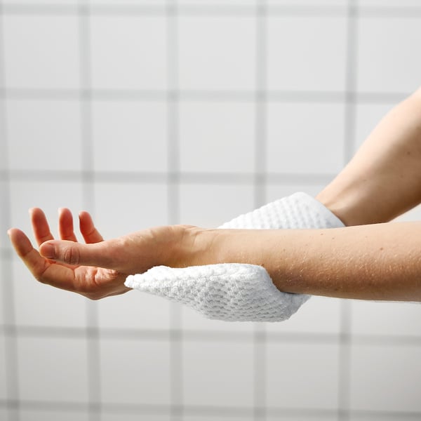 Personne lavant le bras avec une éponge blanche, contre un mur carrelé blanc. Routine d’hygiène simple.