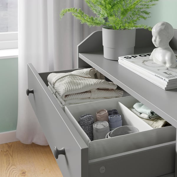 Commode grise moderne avec tiroirs ouverts et fermés, vêtements soigneusement pliés, plante en pot, sculpture et livres.