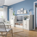 Salon bleu moderne avec mobilier blanc : meuble TV, étagères et chaise. Décoré de plantes et d’objets personnels.