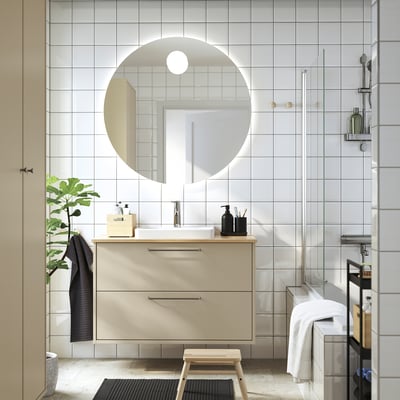 Salle de bain moderne avec grand miroir, armoire beige, lavabo semi-encastré, tabouret en bois, serviette noire, douche en verre, plante.