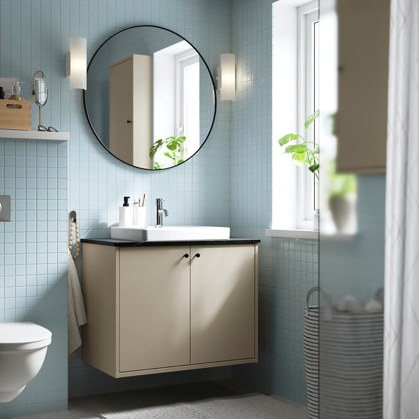 Petit placard de salle de bain beige, fixé au mur, avec un miroir rond, des murs carrelés bleu clair et un lavabo blanc.
