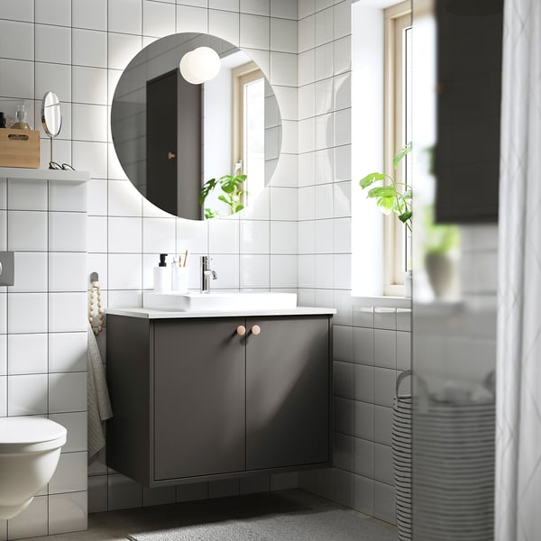 Salle de bain minimaliste moderne : miroir rond, murs carrelés blancs, meuble en HAVBÄCK gris avec lavabo, serviette grise, plante et panier à linge rayé.