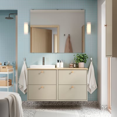 Salle de bains moderne avec carrelage bleu clair, meuble beige HAVBÄCK, lavabo rectangulaire, miroir et deux appliques murales. Deux serviettes blanches sont suspendues à côté de l’armoire.
