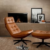 Fauteuil inclinable en cuir marron avec repose-pieds, sur un tapis beige à côté d’une cheminée avec des œuvres d’art et des livres sur le manteau.