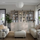 Salon moderne avec des bibliothèques en pin gris clair HAVSTA remplies de livres, un canapé et un pouf blancs, des murs gris et des lampes suspendues.
