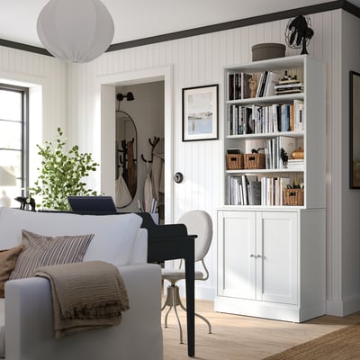 Espace de vie confortable avec étagère en pin HAVSTA, blanc, rempli de livres, décor moderne, canapé gris, bureau noir, plante et art.