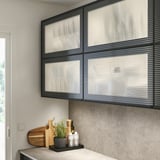 Scène de cuisine moderne avec une grande armoire HEJSTA noire, avec des panneaux de verre teintés foncés, deux fois plus hauts que les articles à proximité, offrant un rangement élégant.