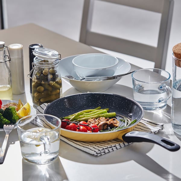 Mise en place de la table avec poêle à frire HEMLAGAD remplie de légumes et de viande, accompagnée de bols, de verres et de bocaux.
