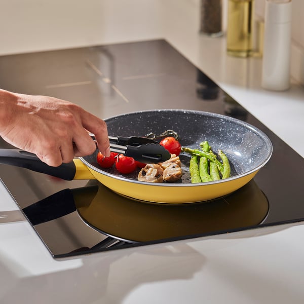 La main jette les ingrédients dans une poêle à frire jaune antiadhésive sur une cuisinière, mettant en valeur une répartition uniforme de la chaleur et un soulèvement facile grâce à sa poignée ergonomique.