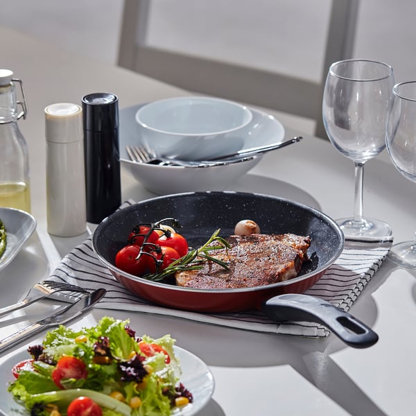 Table dressée avec poêle à frire, assiettes, verres, salade et ustensiles. Un plat de viande frite avec des tomates et des herbes se trouve dans la poêle.