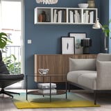 Salon confortable avec tapis coloré, canapé gris et chaise noire. Murs bleus, étagère, armoire en bois, table basse en verre, lampe et plantes.