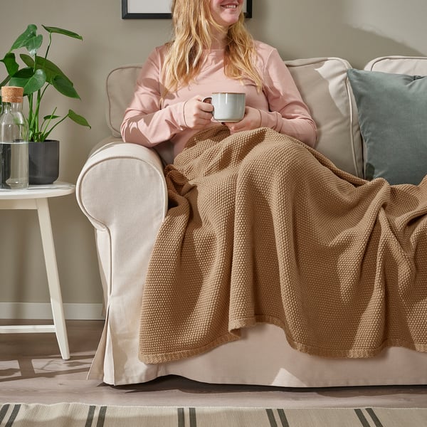 Femme tenant une tasse, enveloppée dans un plaid HUMLEMOTT brun doux sur une chaise beige.