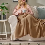 Femme tenant une tasse, enveloppée dans un plaid HUMLEMOTT brun doux sur une chaise beige.