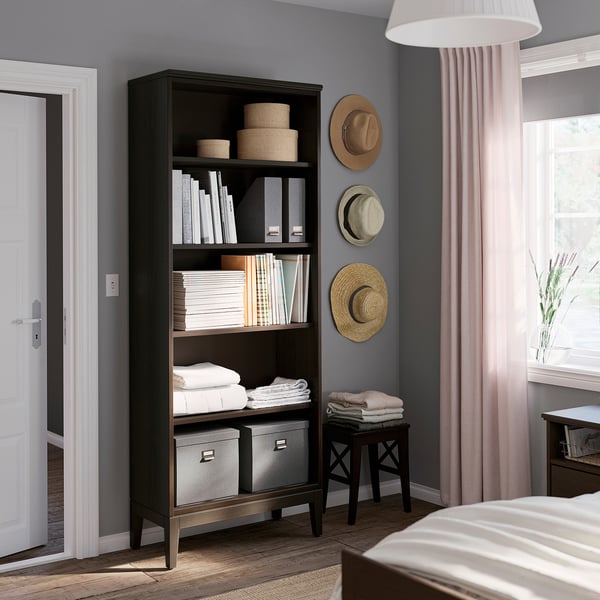 Étagère en bois noir avec de multiples options de rangement, contenant des livres, des paniers et des draps pliés, à côté d’une fenêtre avec des rideaux transparents.