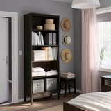 Étagère en bois noir avec de multiples options de rangement, contenant des livres, des paniers et des draps pliés, à côté d’une fenêtre avec des rideaux transparents.