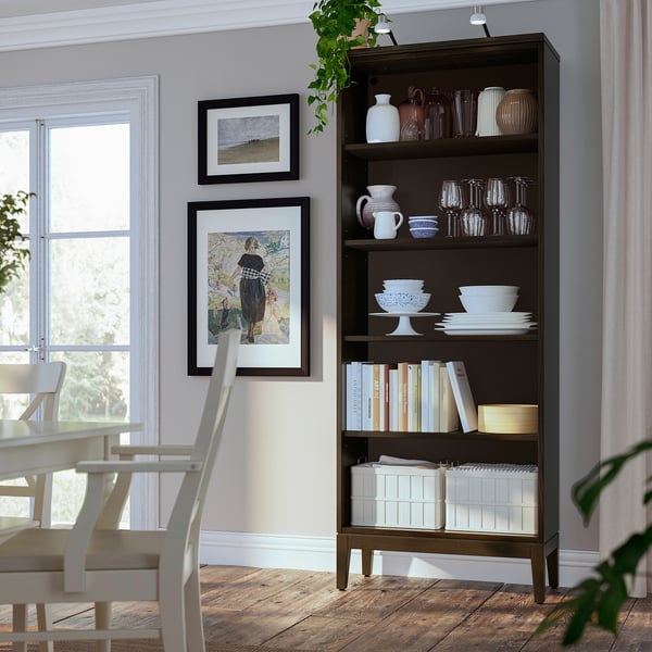 Étagère en bois brun foncé avec divers articles, notamment des bols, des vases et des livres blancs ; à côté des chaises de salle à manger et près de la fenêtre.