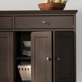 Buffet en bois brun foncé avec des portes pliantes et un tiroir, affichant des assiettes blanches et un panier tressé.